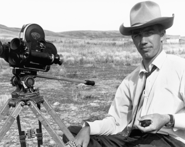 A black and white photograph of diretor Colin Low with a cowboy hat, white shirt, dark tie and pants sitting next to a camera on location during the filming of Director Colin Low on location during the filming of The Days of Whiskey Gap (1961).